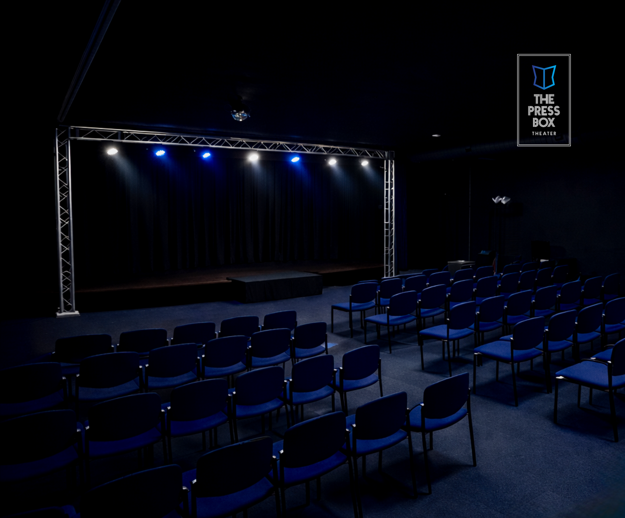 The interior of The Press Box Theater: a warm, intimate 75-seat black box performance space with stage lighting, exposed black ceiling, and close audience seating.